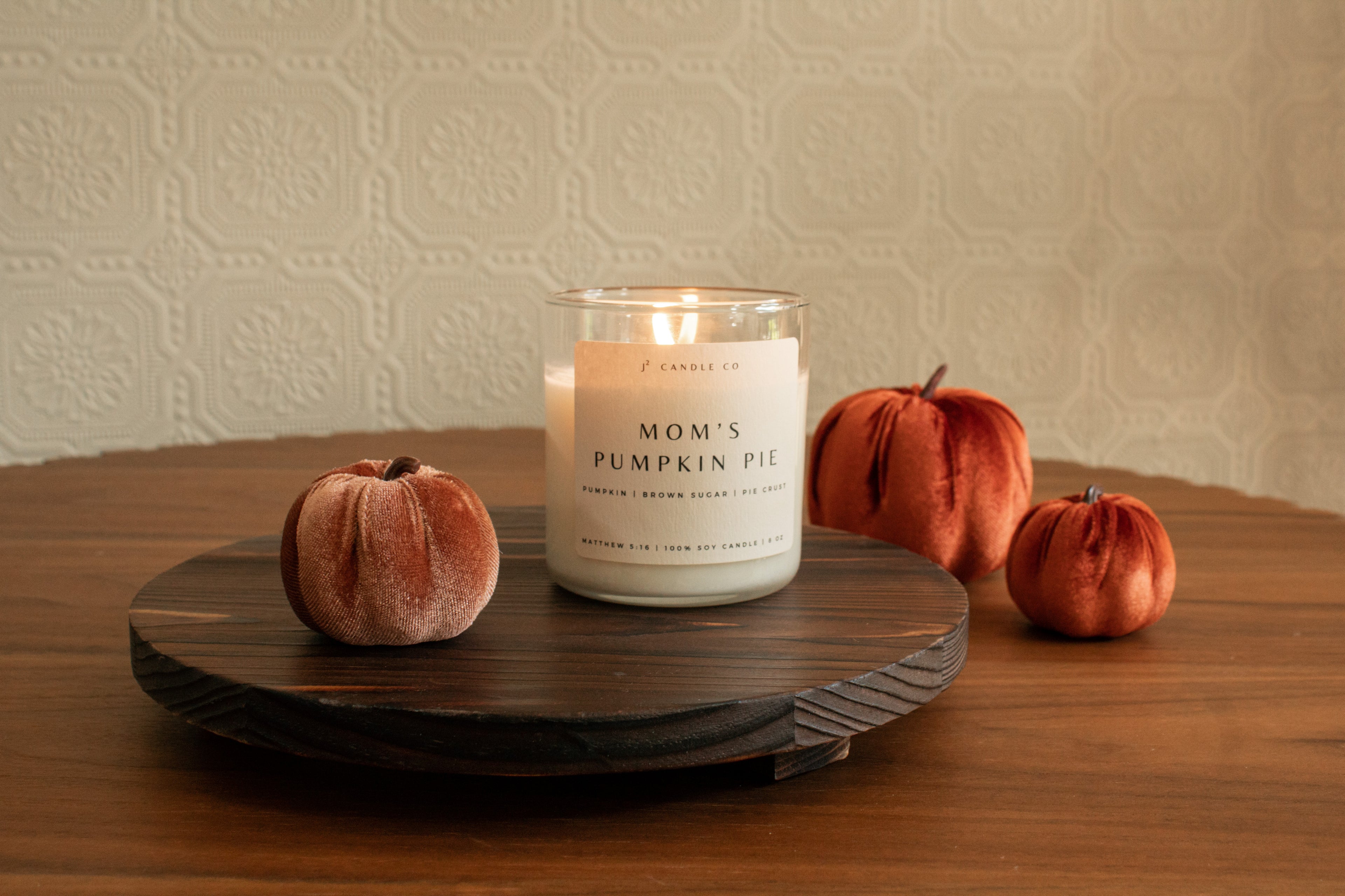 Candle labeled 'Mom's Pumpkin Pie' on a wooden stand with decorative pumpkins on a wooden surface.