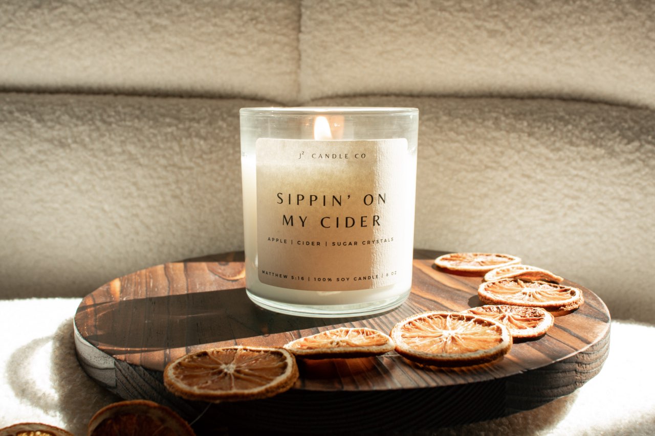 Candle labeled 'Sippin' on my Cider' on a wooden board with dried oranges, set against a neutral background.